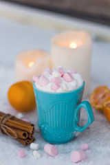 blue mug of drink with whipped cream and marshmallows,candles,dried cinnamon sticks and tangerines on a table covered with snow