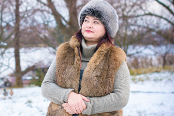 Plus size lady in fur coat wearing hat with snow and nature of Canada in the background. Portrait of smiling plump american woman in grey fur hat. 