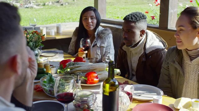 Two women and African man sitting at dinner table with food and wine on it and speaking with friends at party in arbor