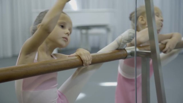 Two little girls do dance exercises on ballet machine. Children in children's studio try to perform complex movements on wooden perlade near mirror.