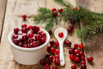 A small bowl of cranberries with sugar on a wooden table with scattered berries and a white spoon © ozhukovaphoto