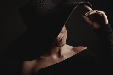 Dark studio portrait of luxury woman in black wide hat and black dress. Hidden eyes.
