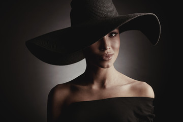 Dark studio portrait of elegant sexy woman in black wide hat and black dress.