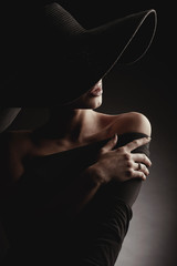 Dark studio portrait of elegant woman in black wide hat and black dress. Hidden half face.