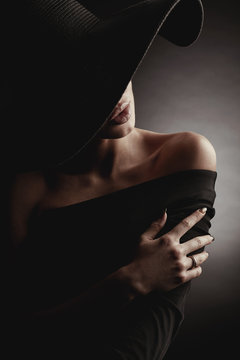 Dark studio portrait of elegant woman in black wide hat and black dress. Hidden half face.