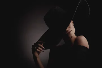 Foto op Plexiglas Dramatic dark studio portrait of elegant woman in black wide hat and black dress. Hidden eyes. © monchak