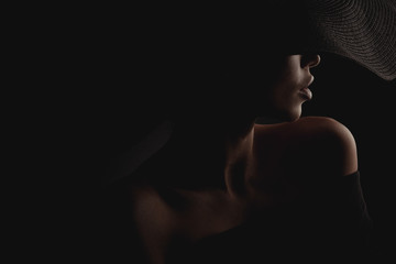 Dramatisches dunkles Studioporträt einer eleganten und sexy Frau in schwarzem, breitem Hut und schwarzem Kleid. Verstecktes halbes Gesicht.