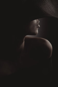 Dramatic dark studio portrait of sexy woman in black wide hat and black dress. Hidden half face.