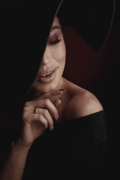 Dramatic dark studio portrait of elegant and sexy smiling woman in black wide hat and black dress.