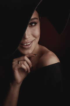 Dramatic dark studio portrait of elegant and sexy smiling woman in black wide hat and black dress.