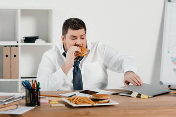 overweight businessman eating hamburger and using laptop in office