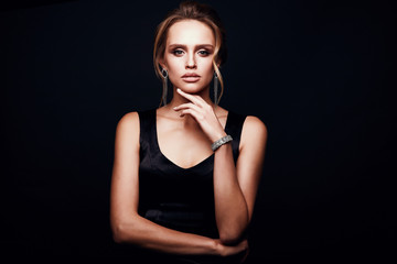 Young amazing blonde sexy woman with glamorous makeup, wearing silky black dress and chic jewelry, posing in studio with dark background