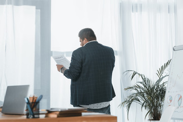 back view of overweight businessman in suit reading document at window in office