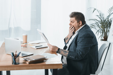 overweight yawning businessman in suit reading newspaper at workplace