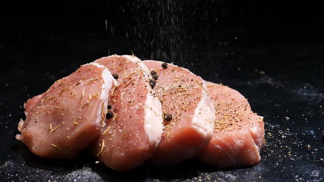Slow motion zooming in shot on four juicy raw pices of pork steaks beeing salted on black board