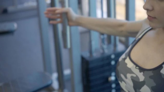 Female athlete is exercising in pec deck machine in gym. Woman sits on seat of iron equipment, holds on to handles and with exhalation, does hand cutting then returns to starting position. Fitness