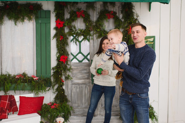 Obraz na płótnie Canvas Happy parents with little son. Child boy in sweater staying on porch snow steps at light house with decorated in red green New Year door at home. Christmas good mood. Family and holiday 2018 concept