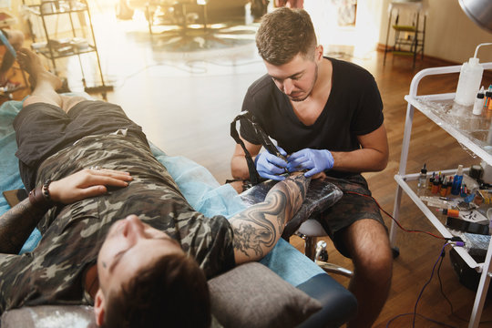 Professional tattooer artist doing picture on hand of man by machine black ink from a jar. Tattoo art on body. Equipment for making tattoo art. Master makes tattooed in light studio.