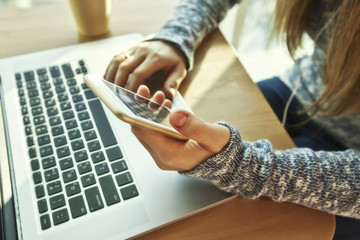 The young woman is sitting at table with modern laptop and uses the phone