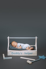 little baby in bodysuit yawning in wooden toolkit with daddys helper lettering and hammer near by