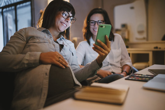 Pregnant woman working in her studio and showing her smartphone to a colleague