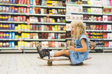 blonde child sitting on skateboard and having fun in supermarket with shelves behind