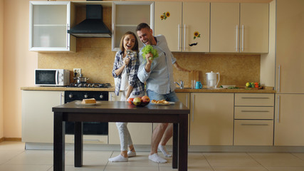 Attractive young joyful couple have fun dancing and singing while cooking in the kitchen at home