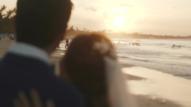 Kissing wedding couple sits on the sea beach watching sunset.
