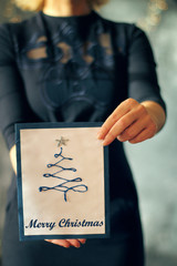 Girl holding a christmas tree card in front of her, face covered, selected focus on the card and hands, Christmas concept