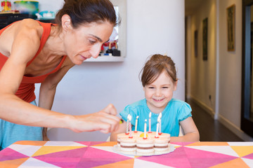 Obraz na płótnie Canvas three years old blonde child smiling watching woman lighting candles with lighter in hand on birthday cake on colorful tablecloth at home 