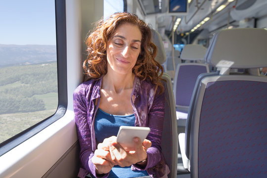 red hair smiling woman dressed in purple and blue, traveling by train sitting, using , surfing internet, reading or typing on mobile smart phone
