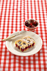 Ein Stück Kirschkuchen auf einer rot-karierten Tischdecke und eine Schale mit frischen Kirschen