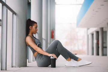 Young fit sporty girl sitting on floor and leaning against the pillar resting after workout.