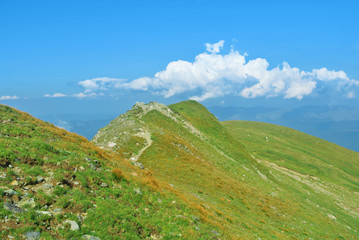 A narrow track road path at mountainside going up around a top of a green peak and a beautiful panoramic view of a chain of mountains at the background on sunny summer day. Tracking in mountains.