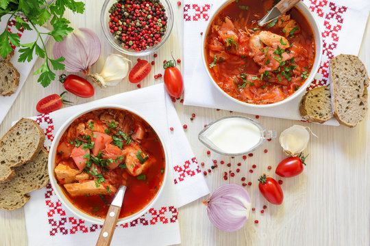 Two bowls of Ukrainian borscht on embroidered napkins, bread, sour cream, spices, onions, garlic, tomatoes and parsley on a white wooden background.