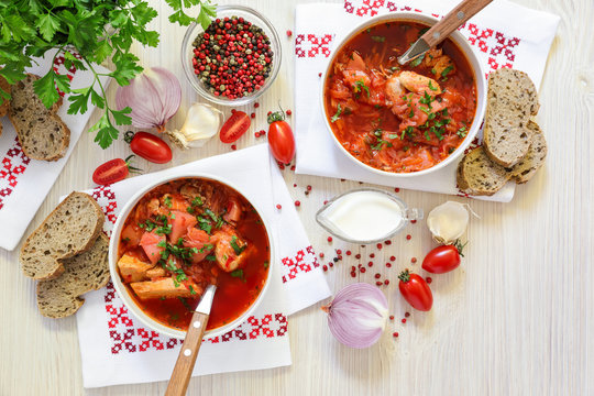 Two bowls of Ukrainian borscht on embroidered napkins, bread, sour cream, spices, onions, garlic, tomatoes and parsley on a white wooden background.