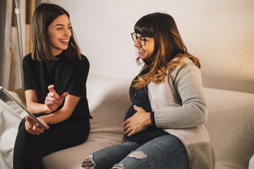 Pregnant woman and her friend look at a tablet on a couch