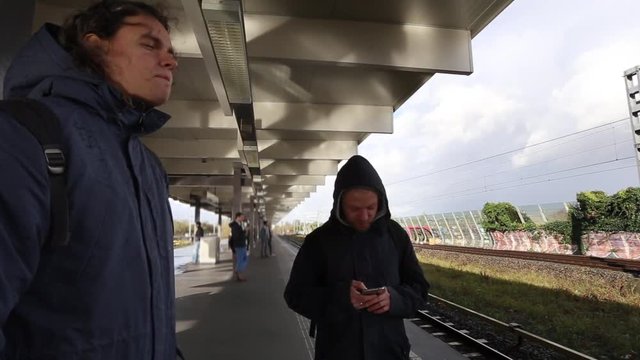 Two men stand on the platform waiting for the train and one of them looks at the phone
