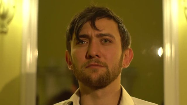 Tired bearded man stands in front of a mirror in the light of lamps, rollback camera
