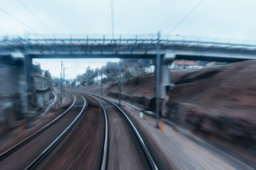 View from the last coach of high-speed suburban train: railway tracks turning to the left under the bridge, houses, poles with wires and dull evening sky; strong motion blur