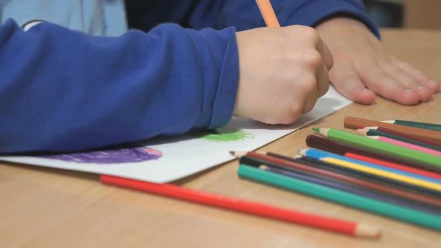 Close-up of hands of unidentified little boy in blue sweater sitting at table drawing pictures using multicolored pencils in kindergarten