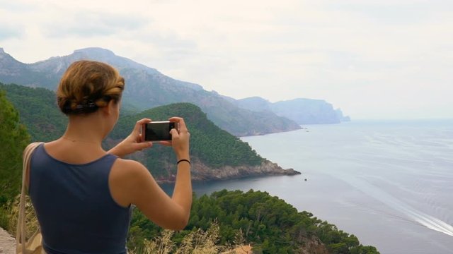 Young happy woman takes smartphone pictures on top of the mountain clifff from the beautiful ocean landscape
