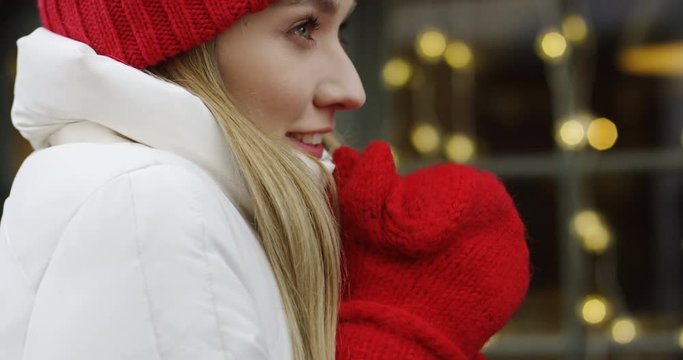 Portrait shot of the beautiful young man in a red hat and gloves standing on the street in winter time and warming his hands and shoulders. Cold day. Christmas lights decorations on the background