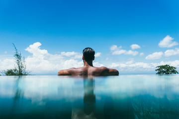 Topless man at infinity pool's edge against sky with fluffy clouds in a hotel