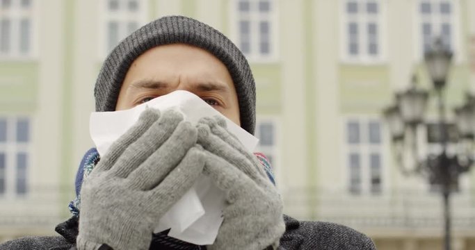 Close up of the ill young caucasian man blowing his nose in the white napkin and sneezing on the town square on the cold winter day. Outdoor