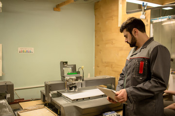 Modern young worker in uniform holding tablet while standing by machine producing layouts