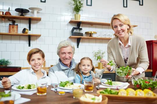 Happy mature female giving fresh vegetable salad to her husband and grandchildren during breakfast