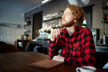 Inspired young man with pencil and notepad thinking of new creative ideas while sitting by table