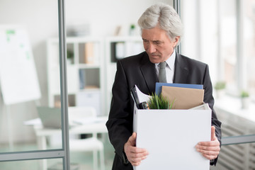 Unhappy mature businessman or director holding box with his things while leaving his office