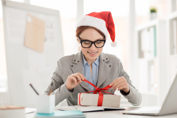 Smiling young impatient employee untying red ribbon on top of package with xmas gift